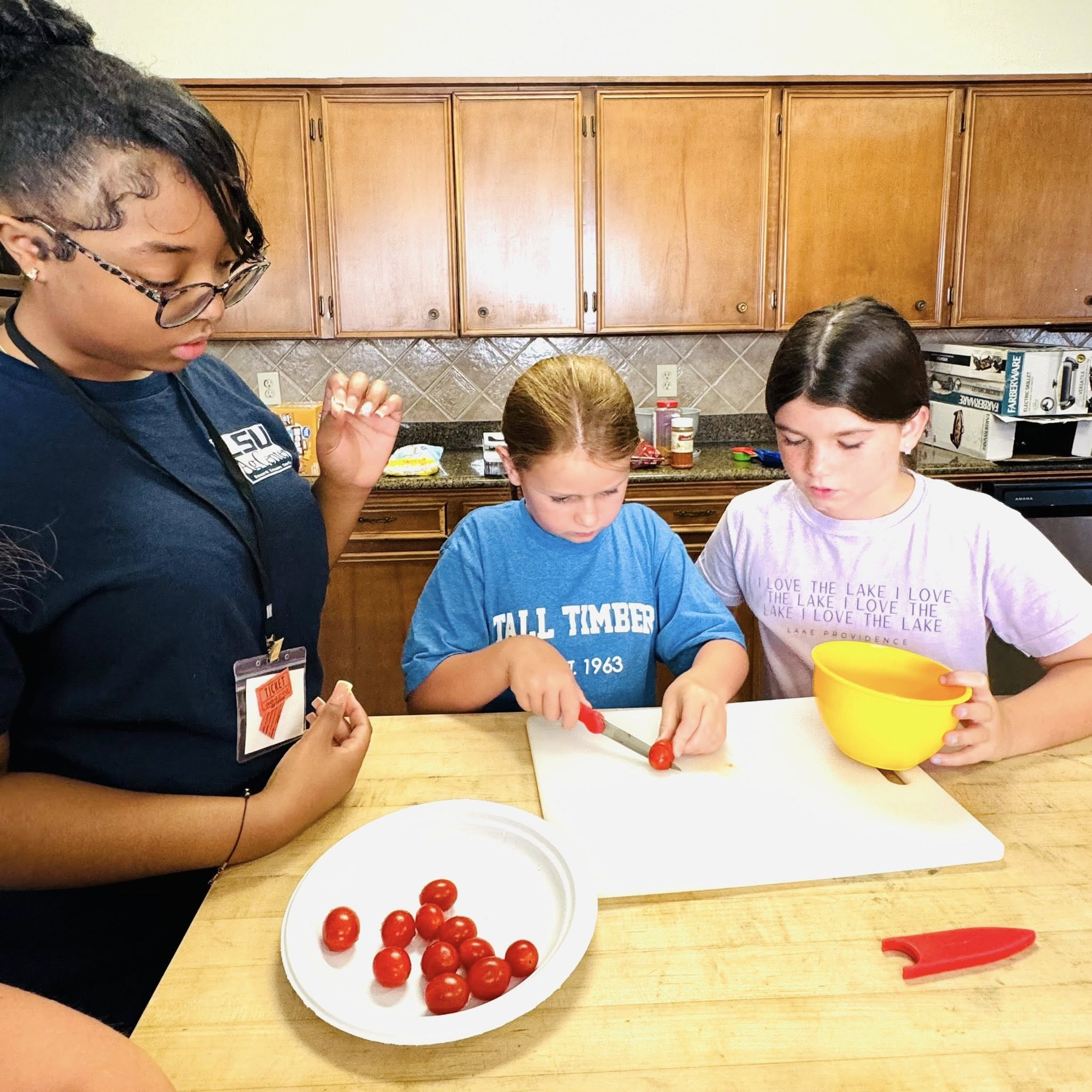Two girls watching another girl slice cherry tomatoes.